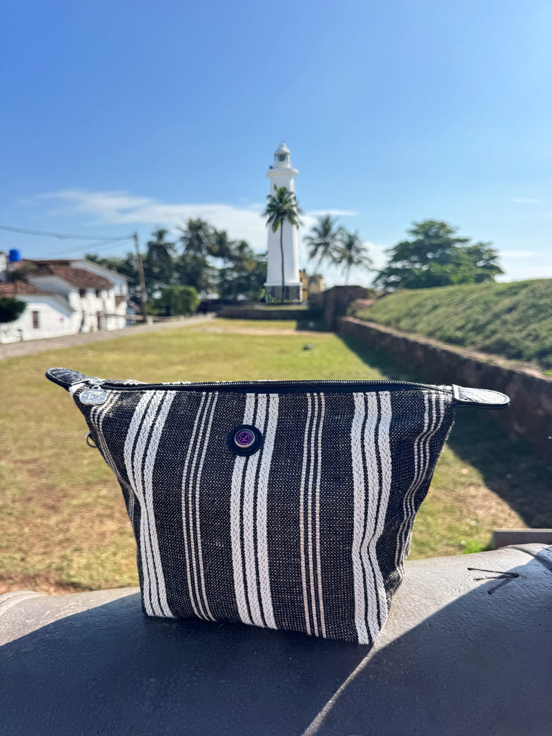 The Toiletry Bag Black & White GALLE rests on a ledge in the foreground, with a white lighthouse, palm trees, grass, and blue sky in the background.