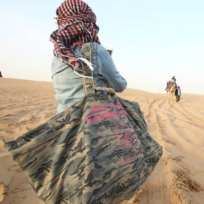 Eine Person, die eine große Tragetasche aus Camouflage-Segeltuch auf Sanddünen trägt, im Hintergrund sind Kamele und Menschen zu sehen.