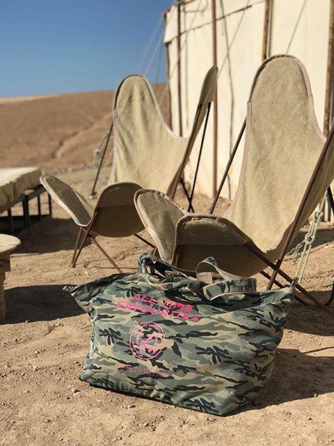 Een middelgrote draagtas van canvas in camouflageprint, geplaatst voor twee stoelen en een beige tent in een woestijnlandschap.