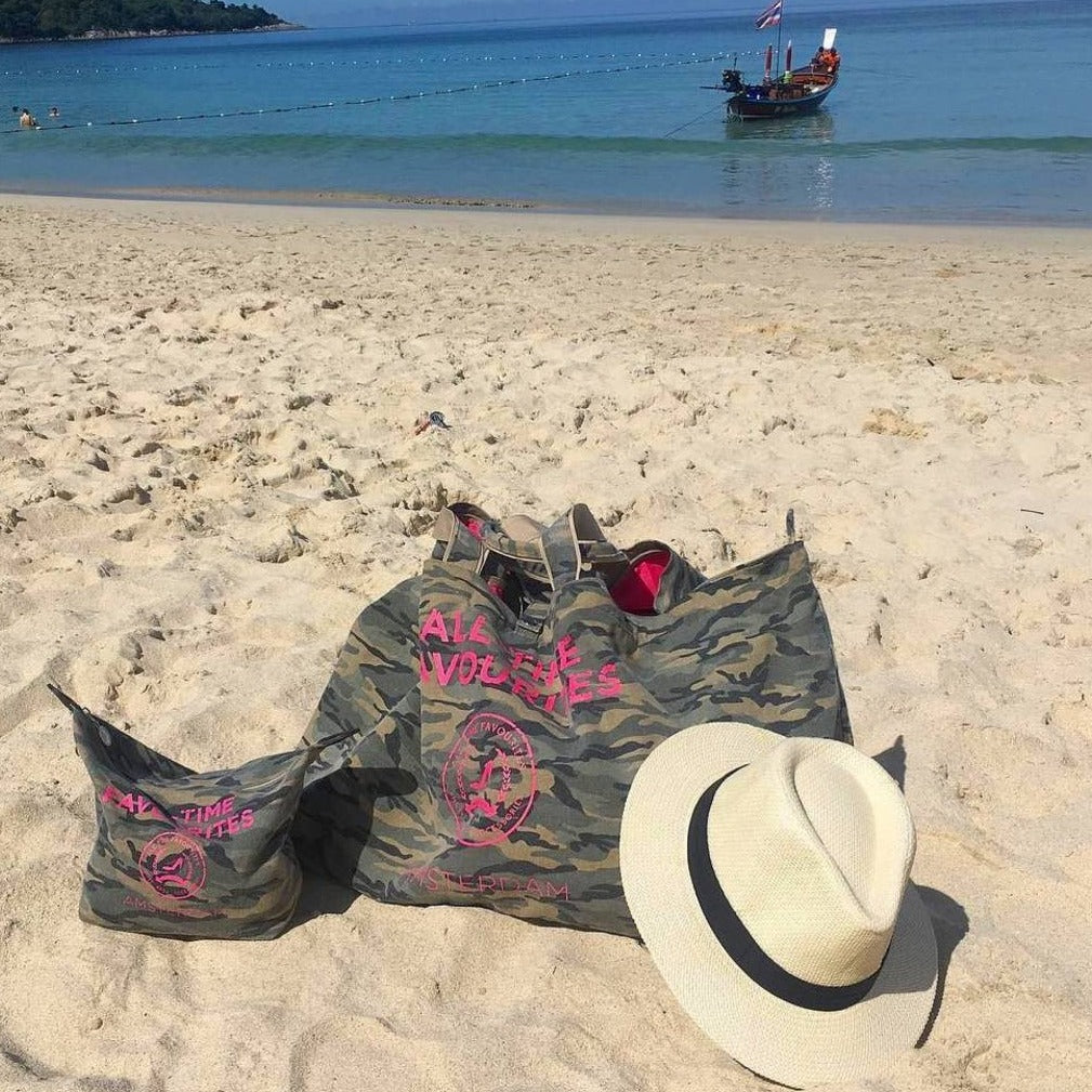 Camouflage-toilettas op een zandstrand met een strohoed, een kalme zee, een boot in de verte en een heldere hemel.